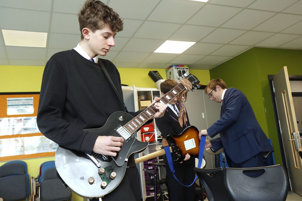 In the music room two youths practice with their electric guitars attended by the assistance of the music teacher