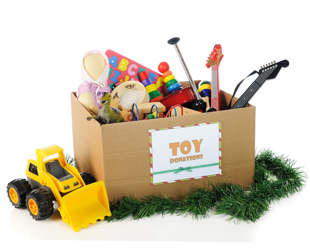 A large corrugated box with a "Toy Donations" sign. Its filled with assorted toys and surrounded by Christmas garland and a toy bulldozer. On a white background.